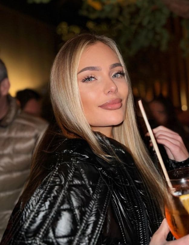 Meriem stands in a dim indoor setting wearing a black quilted puffer jacket, holding a drink with a straw, with blurred people in the background.