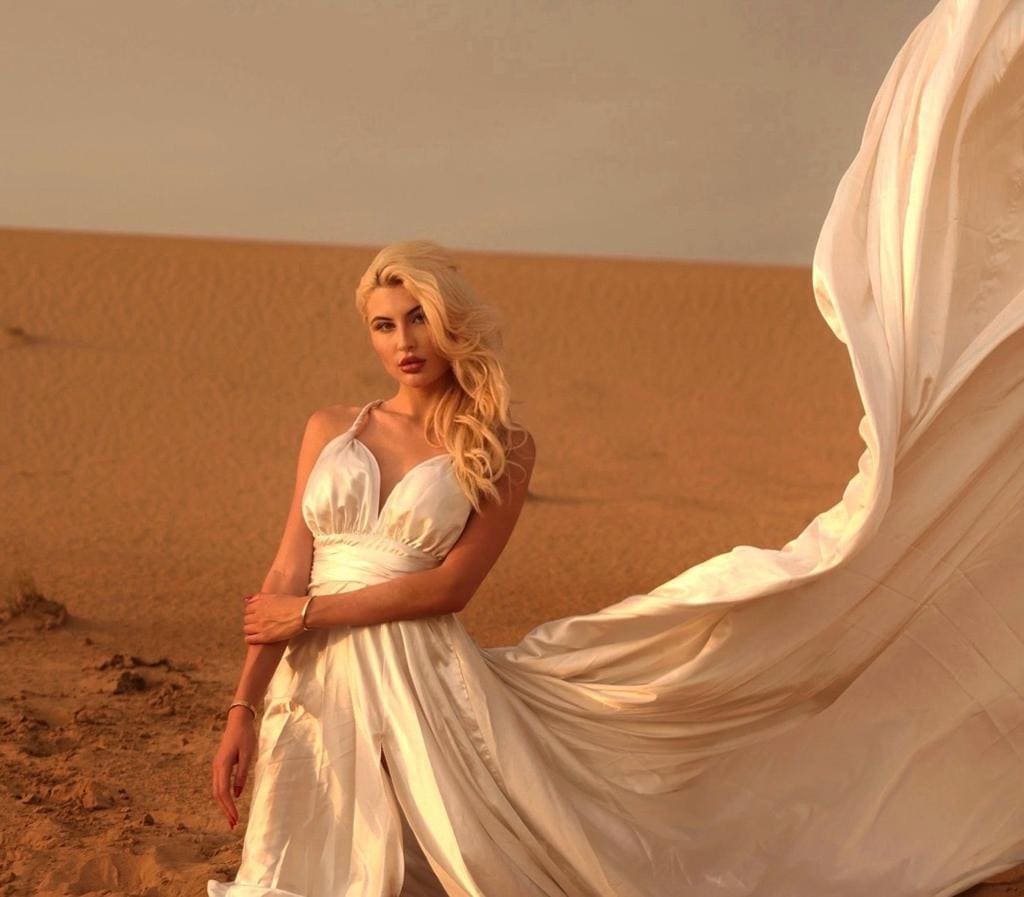 A blonde woman in a white satin dress stands in a desert with arms crossed as white fabric billows behind.
