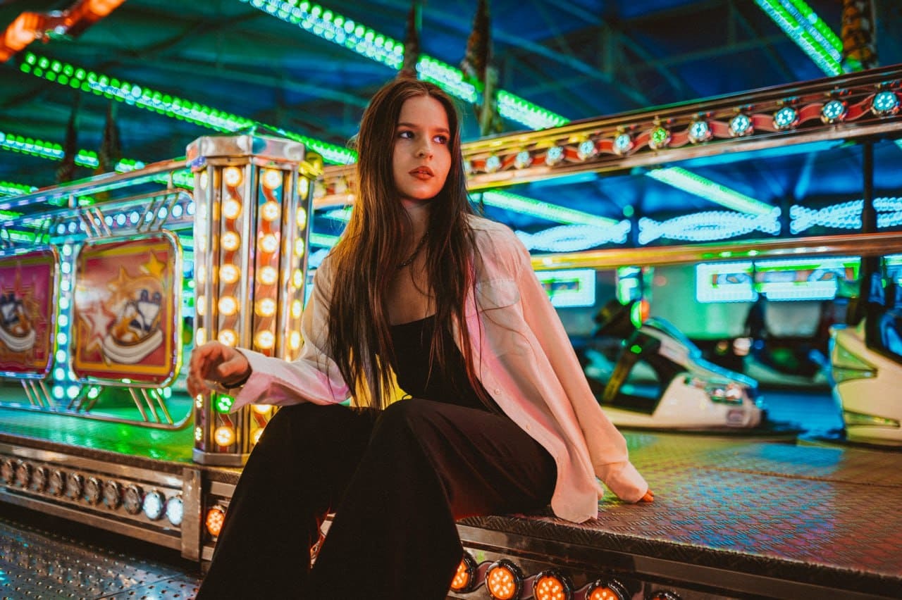 A woman sits on an arcade platform in an open white shirt over a black top, surrounded by neon lights and game machines.