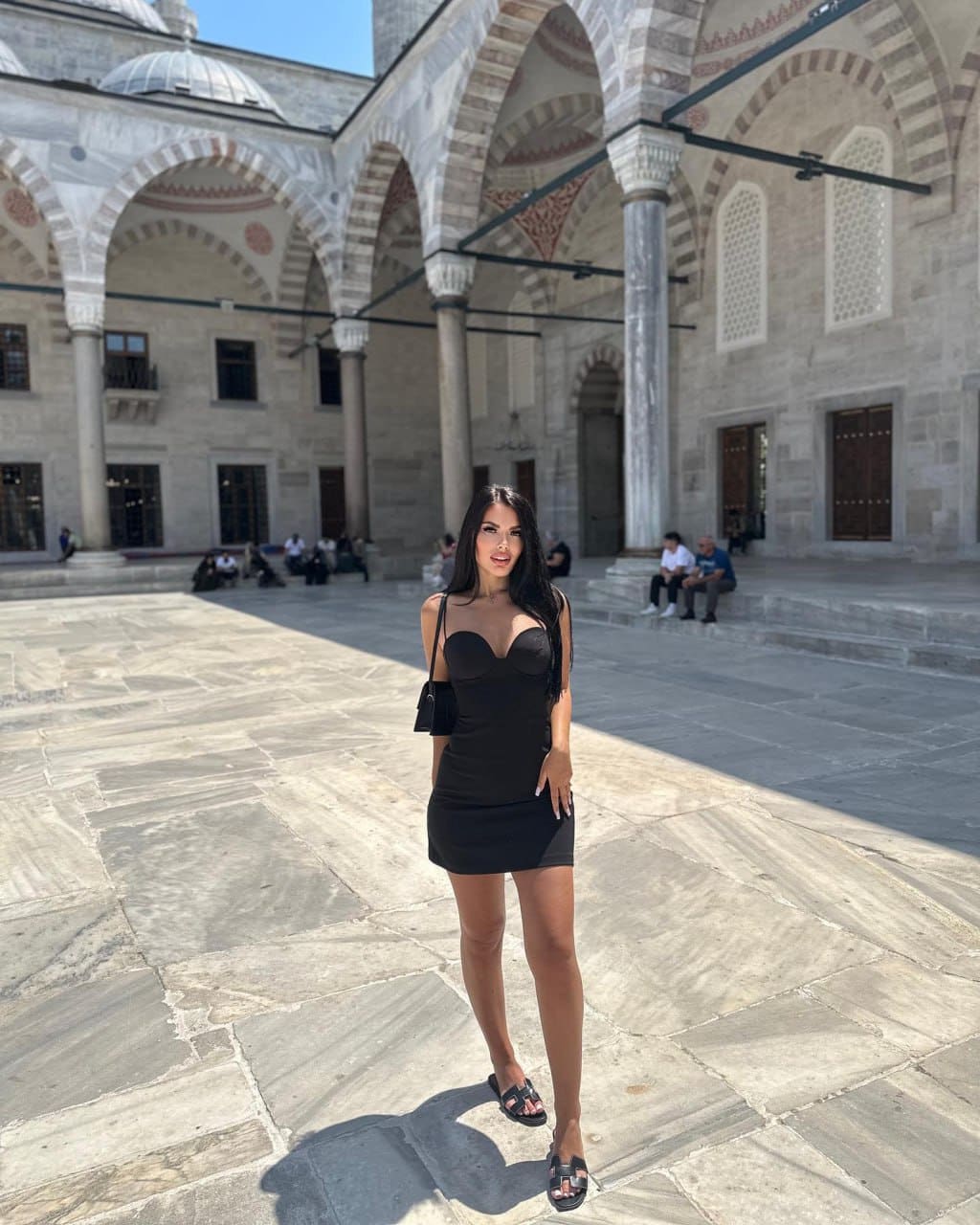 A woman stands in a stone courtyard wearing a short black dress and sandals, holding a small bag by an arched building.