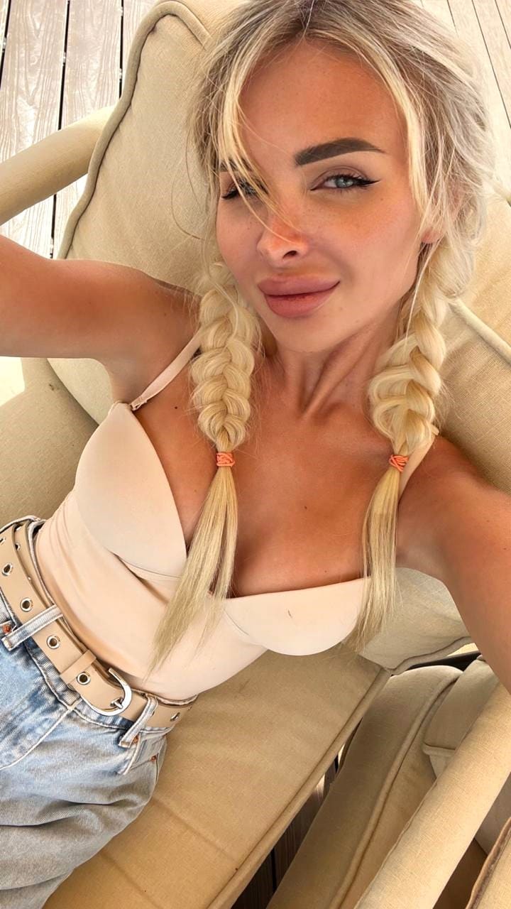 A blonde woman with braids sits on a beige lounge chair wearing a cream strapless top and denim jeans, posing with one arm raised outdoors.