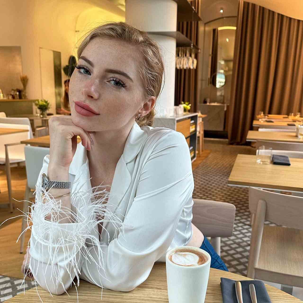 Sonya sits at a wooden restaurant table in a white feather-trimmed blouse, resting her chin on her hand beside a coffee cup.