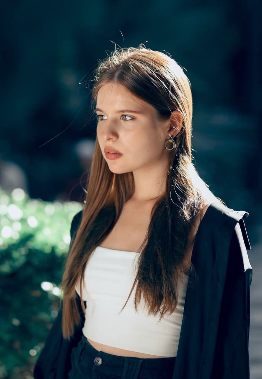 A woman stands outdoors in a white cropped tank and dark open jacket, looking aside against a blurred green background.
