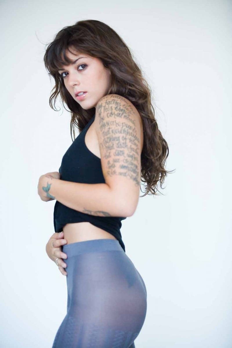 Nova stands in profile against a white background, wearing a black cropped tank and sheer grey leggings, looking over her shoulder.