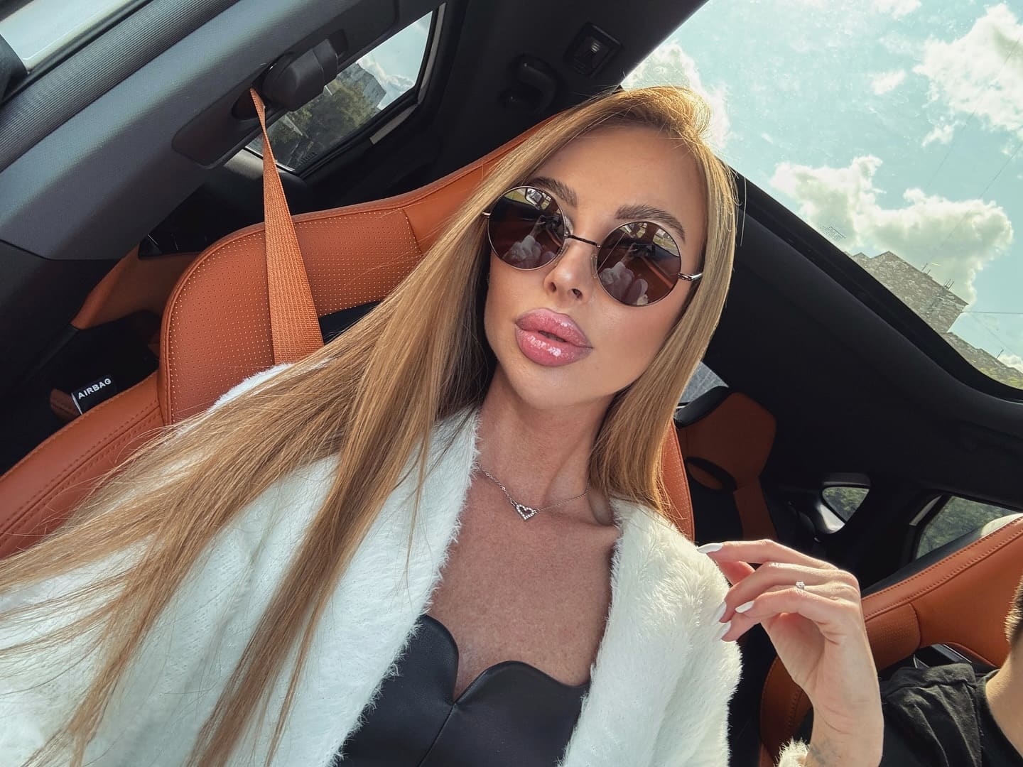 Woman with sunglasses sits in a car driver's seat wearing a white fuzzy cardigan and heart necklace, one hand raised near her shoulder.