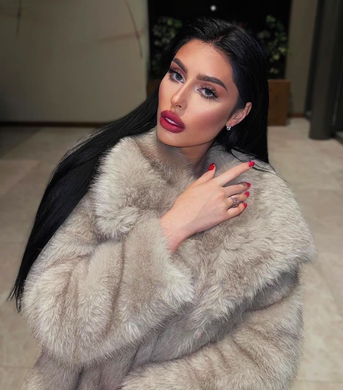 Aleks sits indoors in a thick grey fur coat, raising a hand with red nails and a ring to her chest.