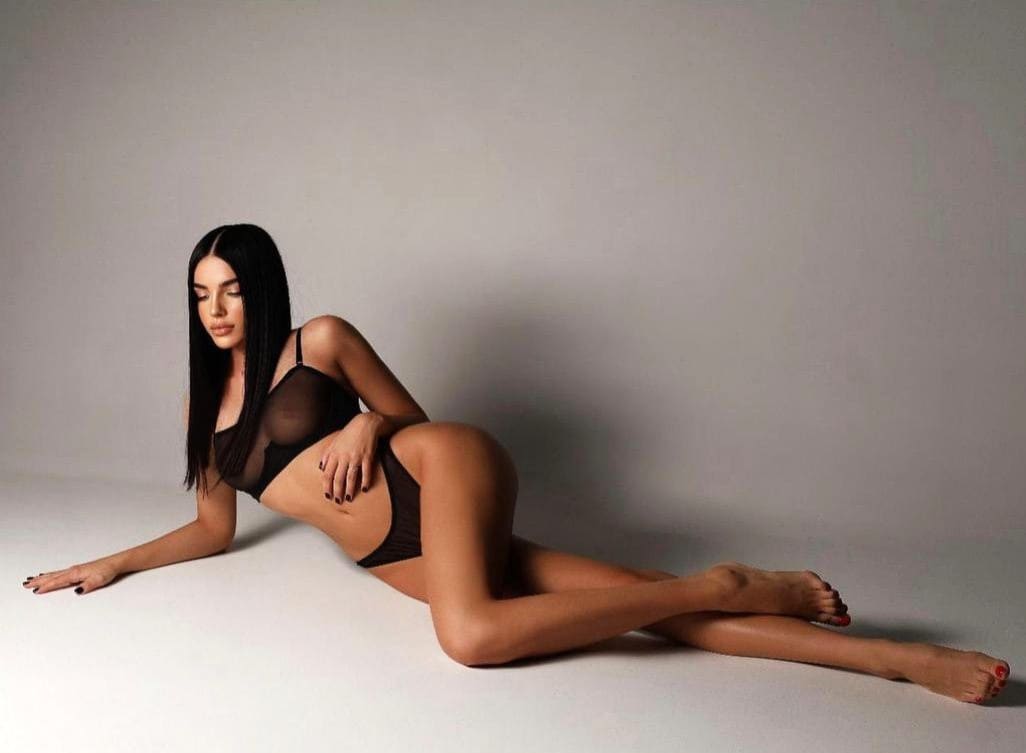 Lying on her side on a white studio floor, a woman in a sheer black bra and briefs extends one arm forward while resting the other on her hip.