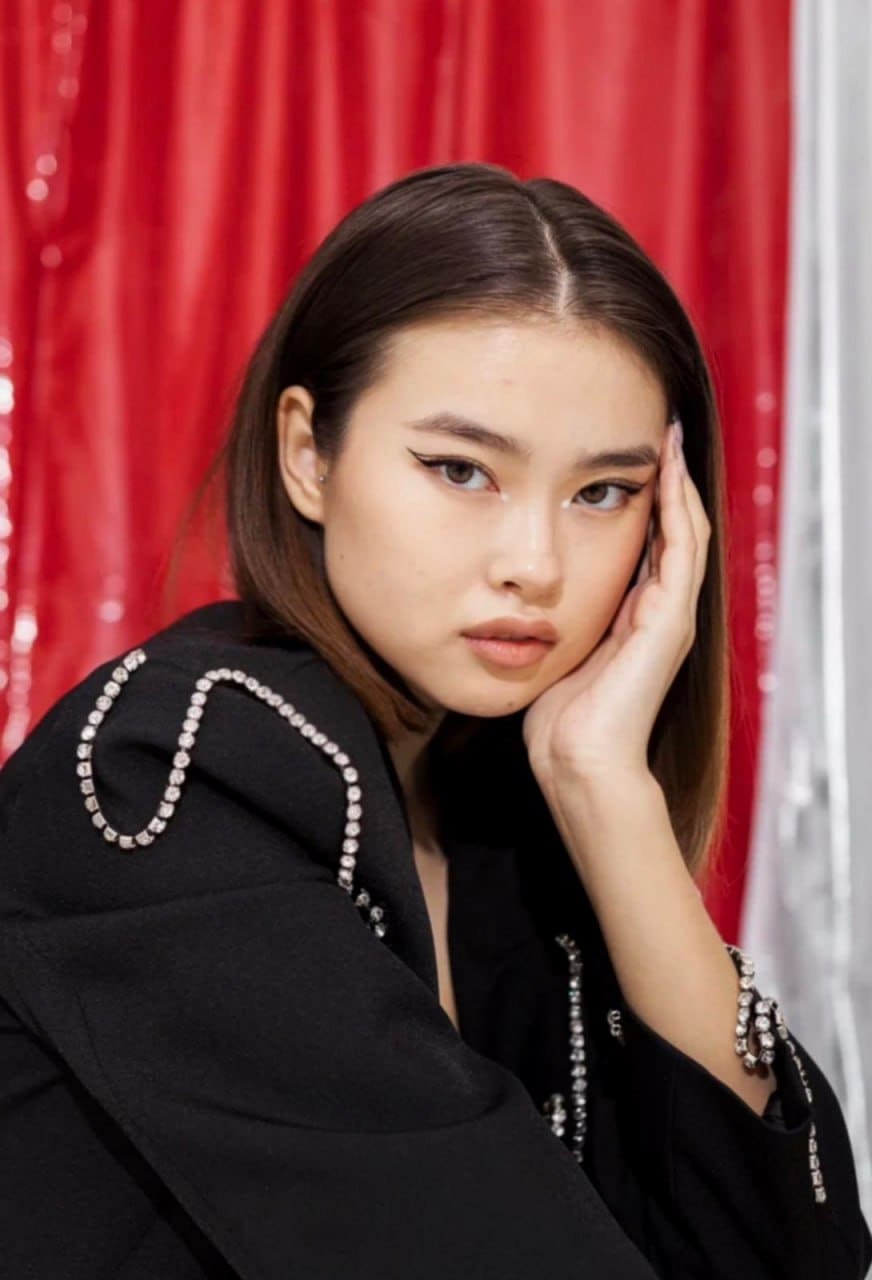 A woman in a black jacket with crystal trim rests her chin on her hand against a red curtain backdrop.