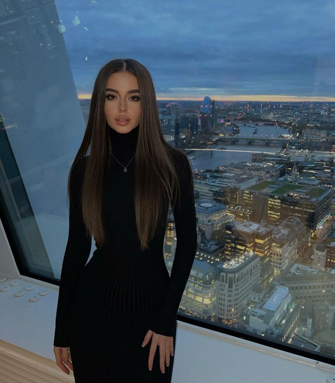 Maria stands in a fitted black turtleneck dress with one hand on her hip, positioned before a window overlooking a city skyline at dusk.
