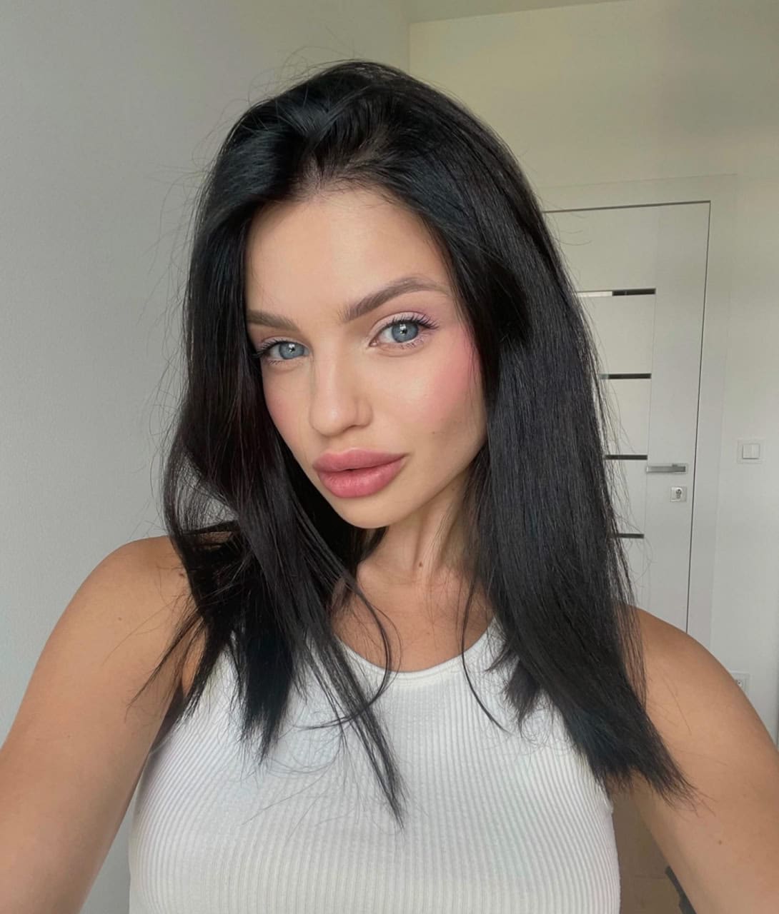 Vladislava stands indoors in a white ribbed tank top, facing the camera with long dark hair and blue eyes against a white door.