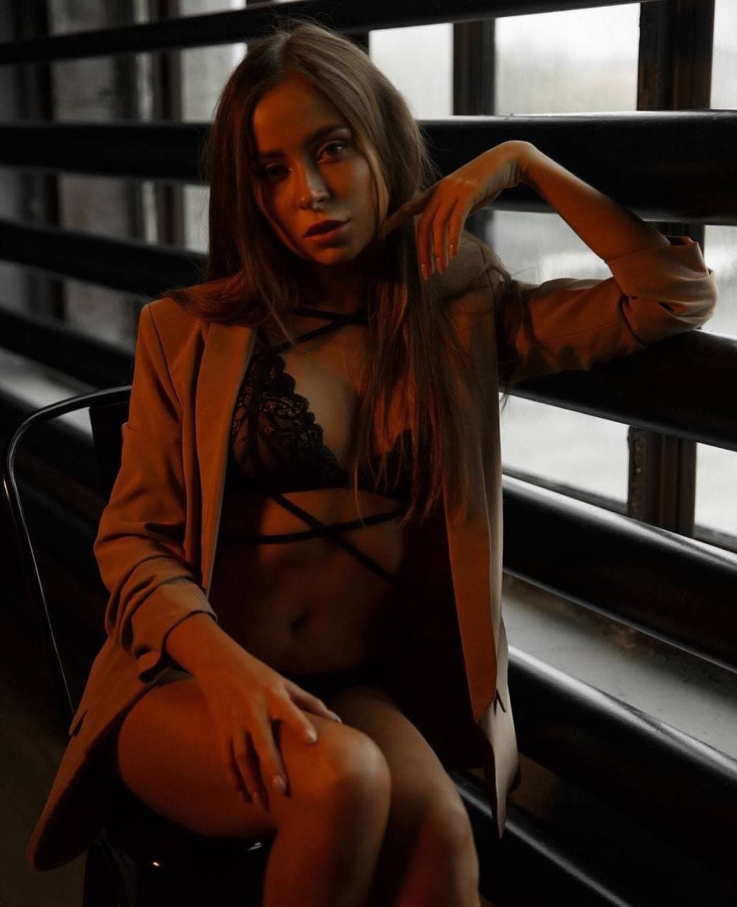 A woman sits on a chair wearing an open tan blazer over a black lace bralette, resting her arm on a dark metal railing.