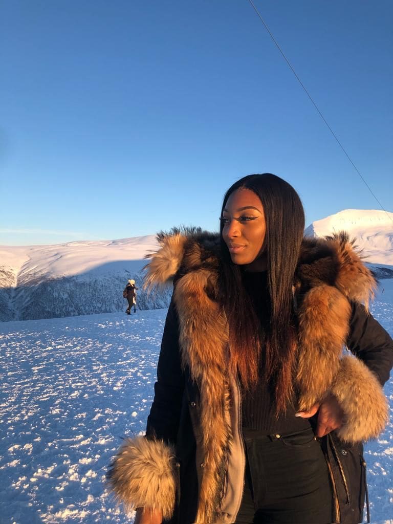 A woman in a black coat with fur collar stands in a snowy mountain landscape under a blue sky.