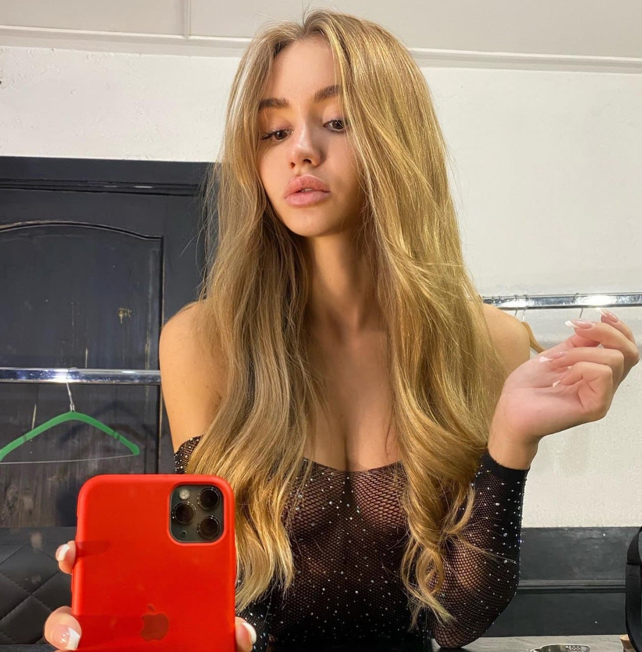 Josetta takes a mirror selfie in a black off-the-shoulder mesh top with sparkly dots, holding a red phone in a room with a dark cabinet.