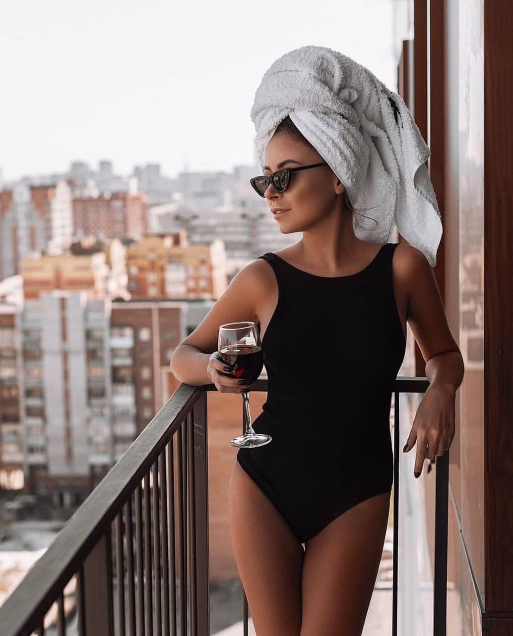 A woman in a black one-piece swimsuit and sunglasses holds a glass of red wine on a balcony with city buildings behind her.