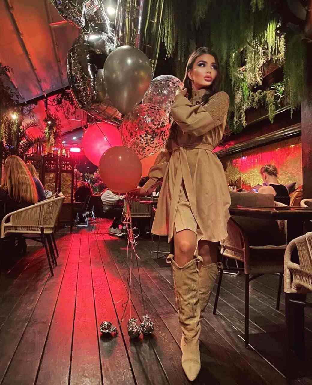 A woman stands on a wooden deck in a dim restaurant, wearing a tan belted dress and suede boots, holding red and silver balloons.