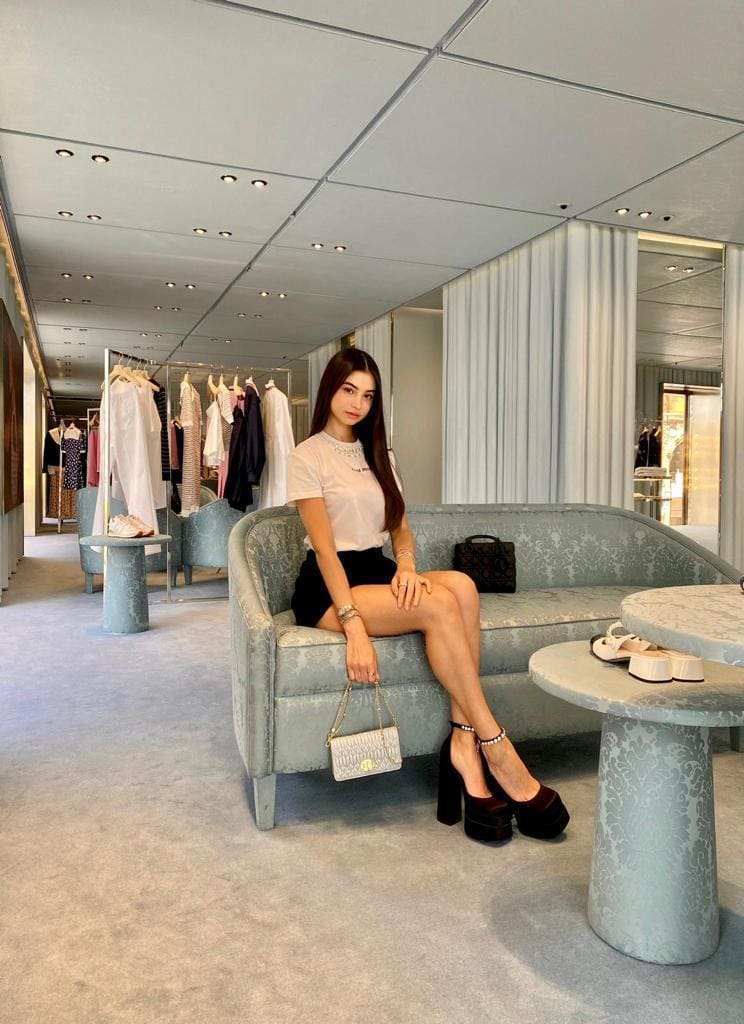 A woman sits on a blue patterned sofa in a store, wearing a white t-shirt, black mini skirt, and platform heels.