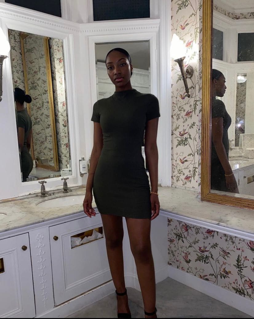 A woman in an olive-green mini dress and black heels stands in a bathroom with floral wallpaper.