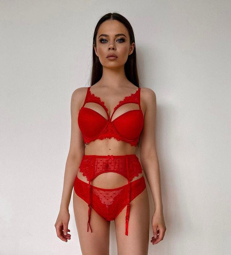 Diona stands facing the camera in red lace lingerie with cutout panels against a plain light wall.