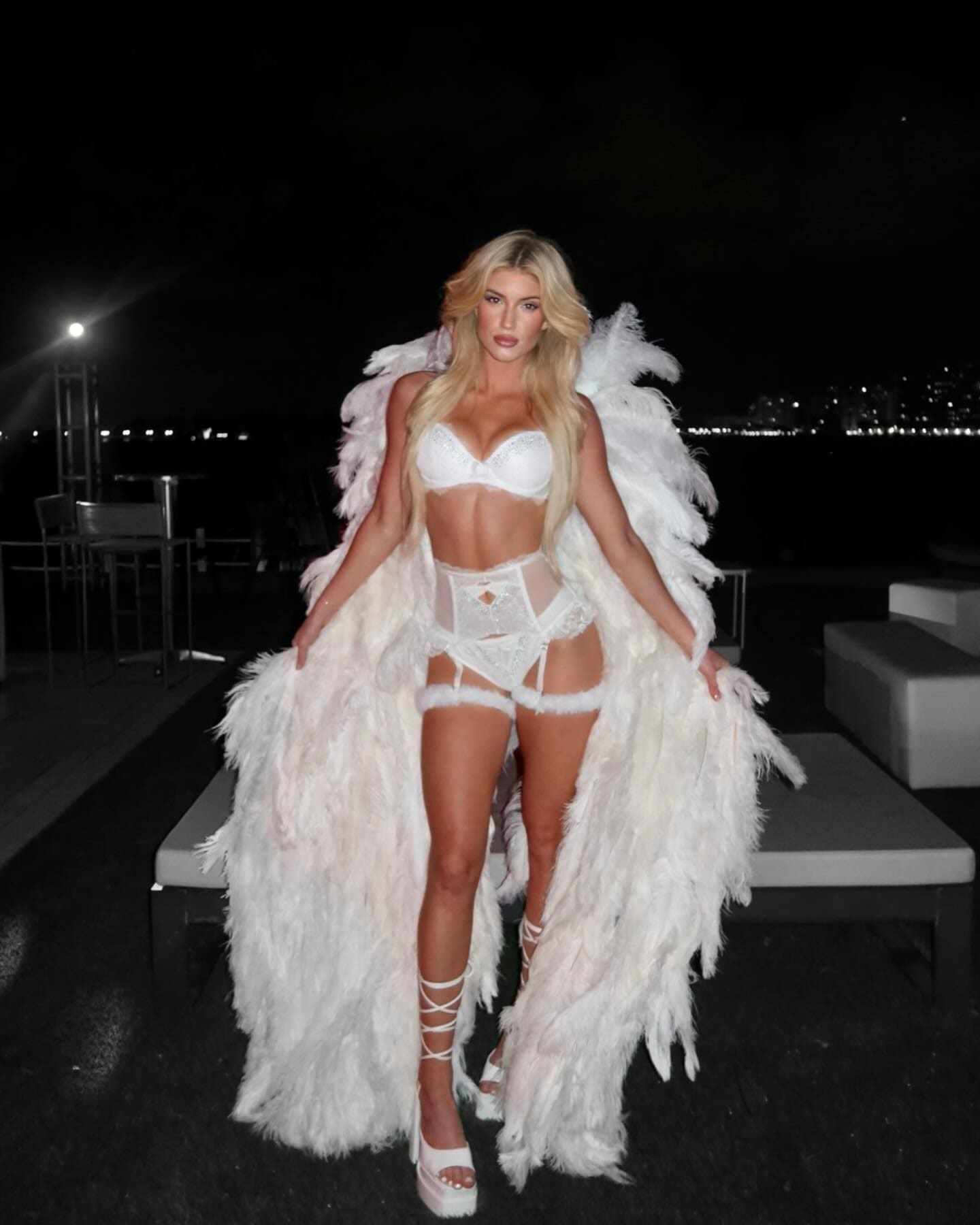 A blonde woman in a white lace bodysuit and feathered coat stands outdoors at night, wearing strappy white platform heels.
