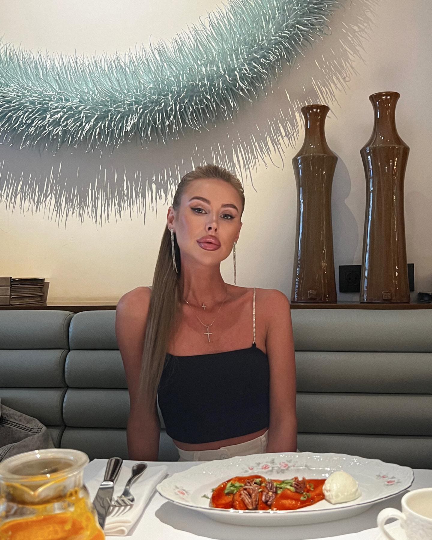Tamilа sits at a restaurant table in a black crop top and white bottoms, with pasta and juice before her.