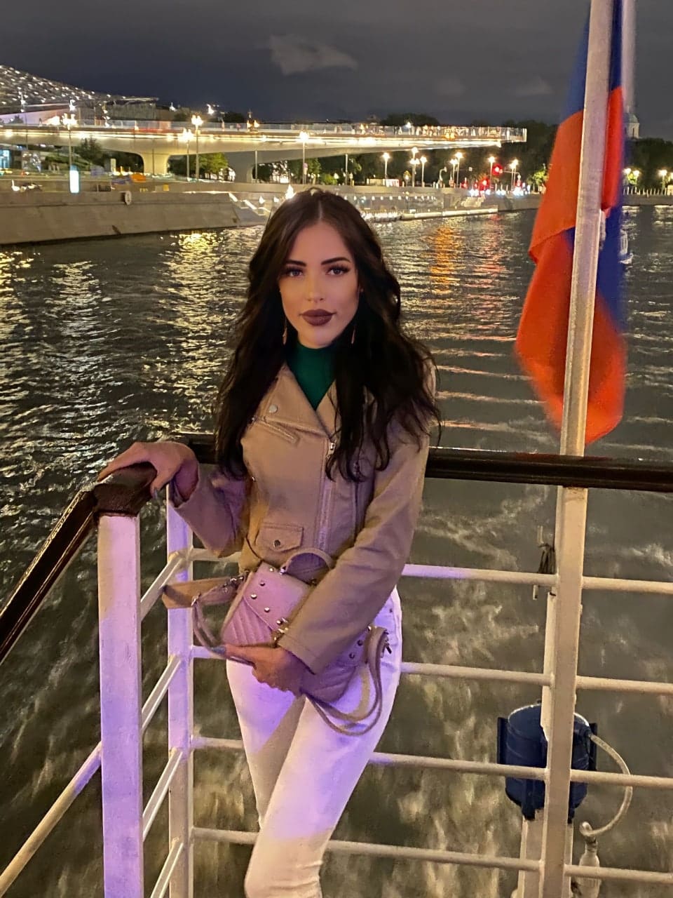 Berta stands on a boat deck at night in a tan leather jacket, green turtleneck, and white pants, leaning on the railing.