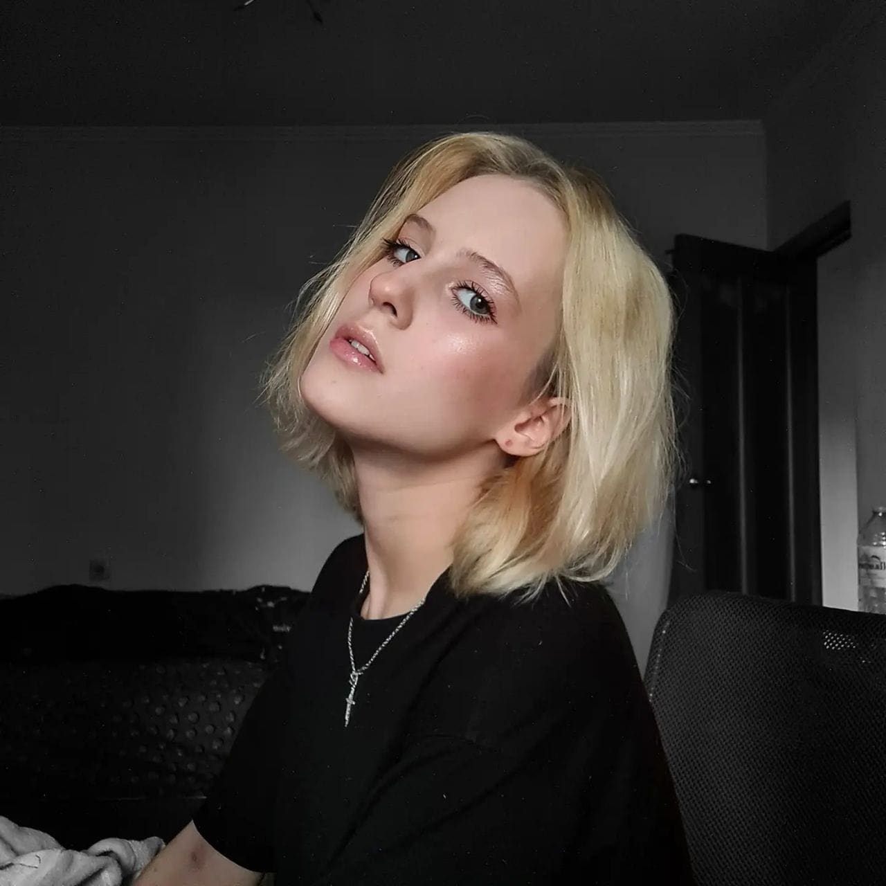 A blonde woman with a bob haircut tilts her head back in a black t-shirt and cross necklace, seated in a dimly lit room with dark furniture.