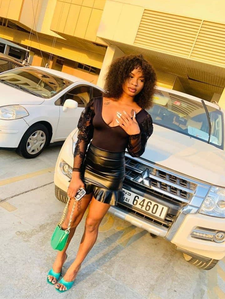 A woman with curly hair poses before a white SUV in a parking lot, wearing a black lace top, leather skirt, and turquoise heels, holding a green bag.