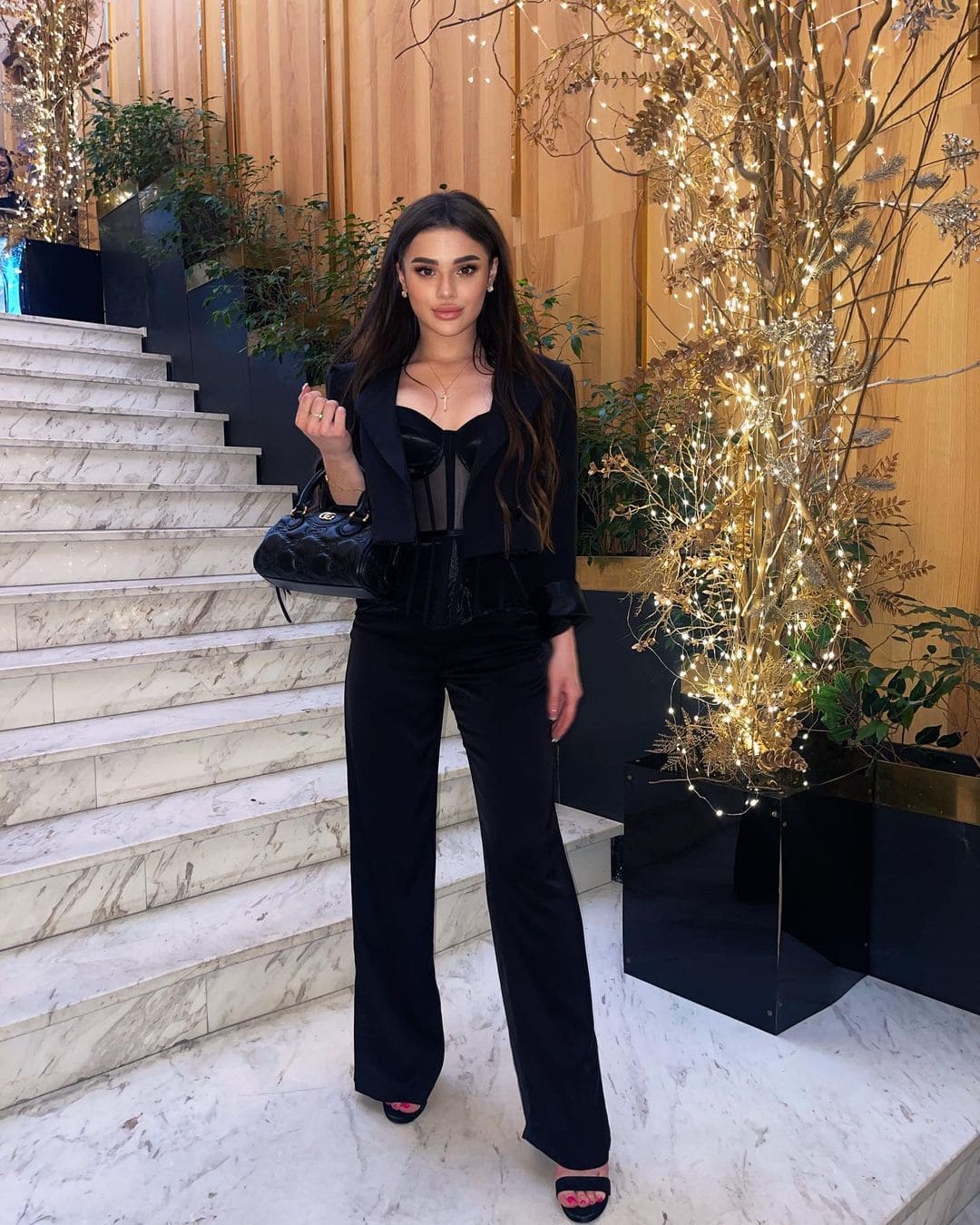 Albina stands on a marble landing in a black cropped blazer, sheer top, wide-leg trousers, and heels, holding a quilted handbag near a lit tree.