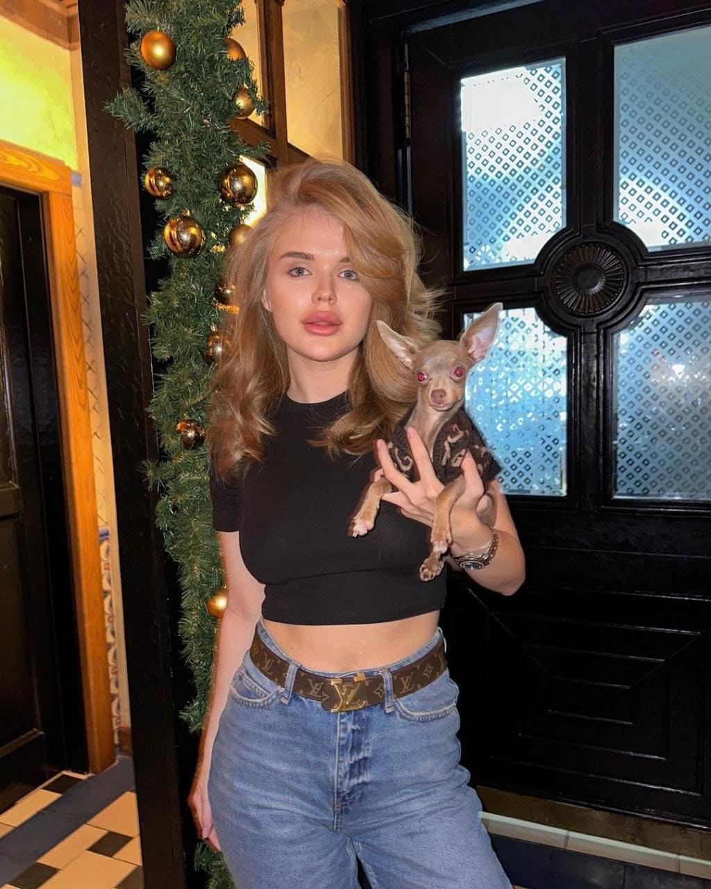 A woman in a black crop top and jeans holds a small tan dog indoors near a Christmas garland.
