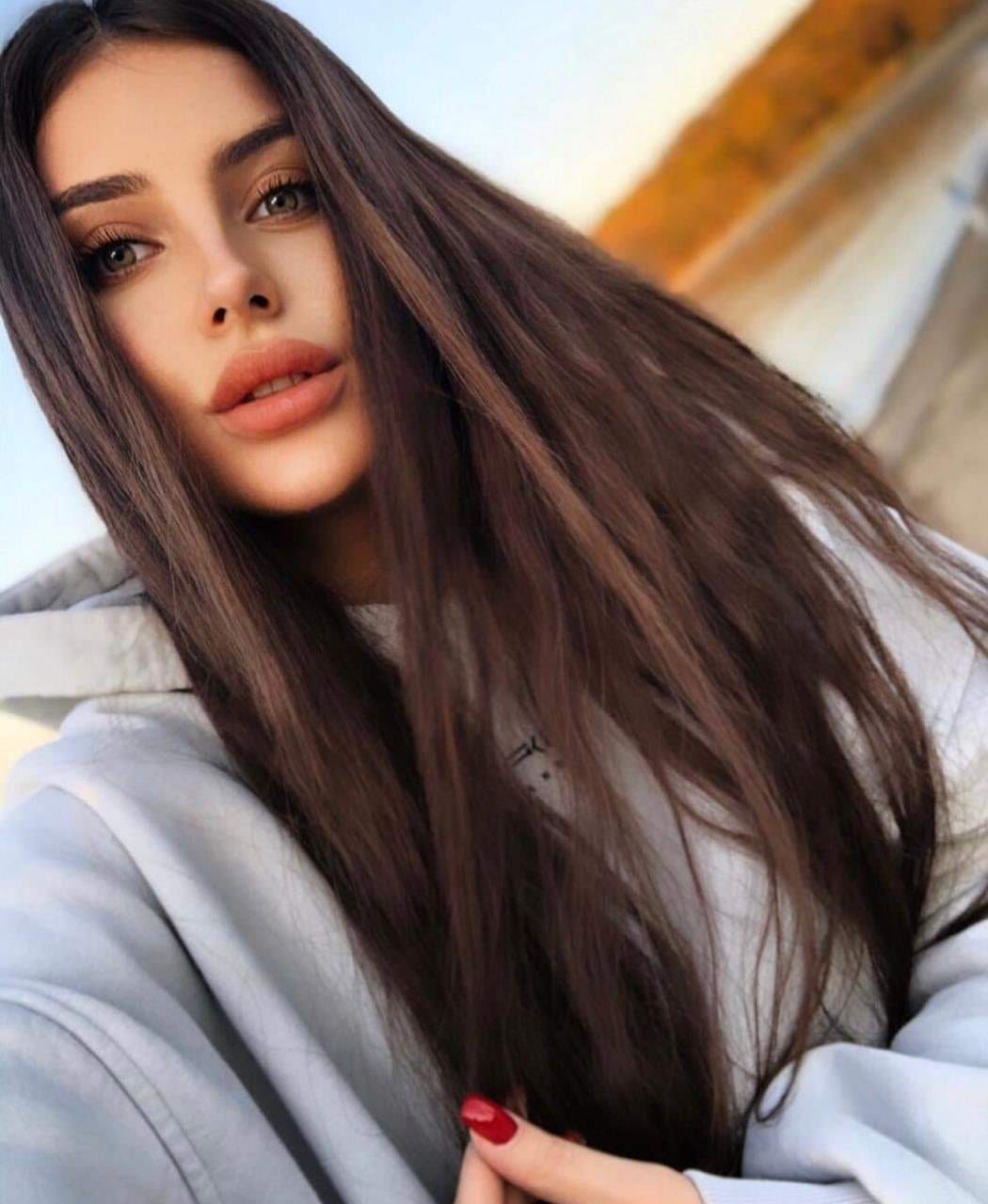 Close-up of a woman in a grey hoodie with red nails, hand raised near her chest, against a blurred outdoor background with warm tones.