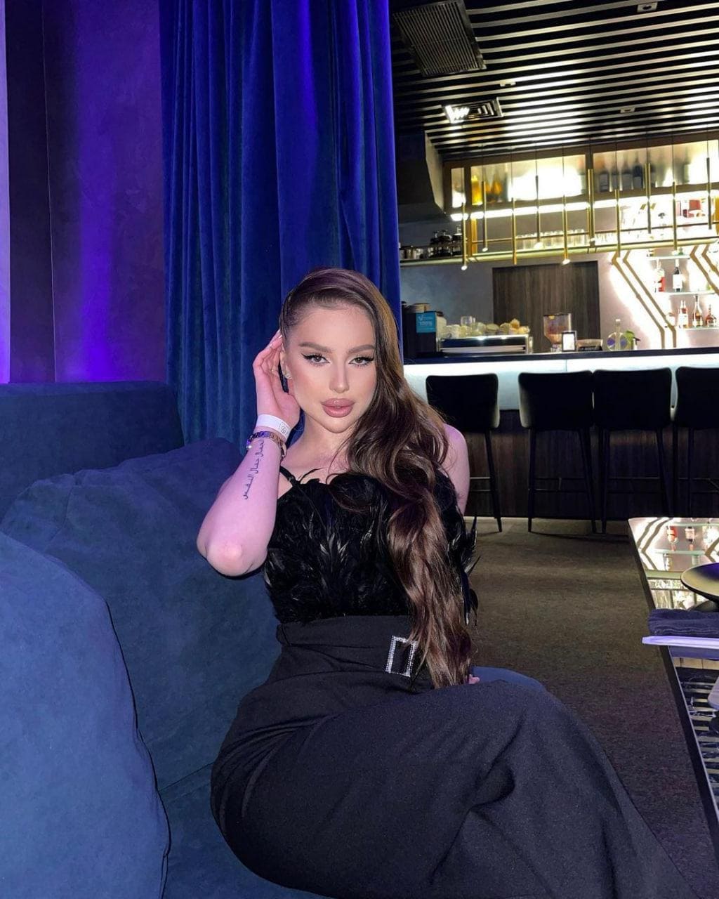 A woman in a black ruffled top and long skirt sits on a blue couch, one hand raised to her head in a dim lounge.