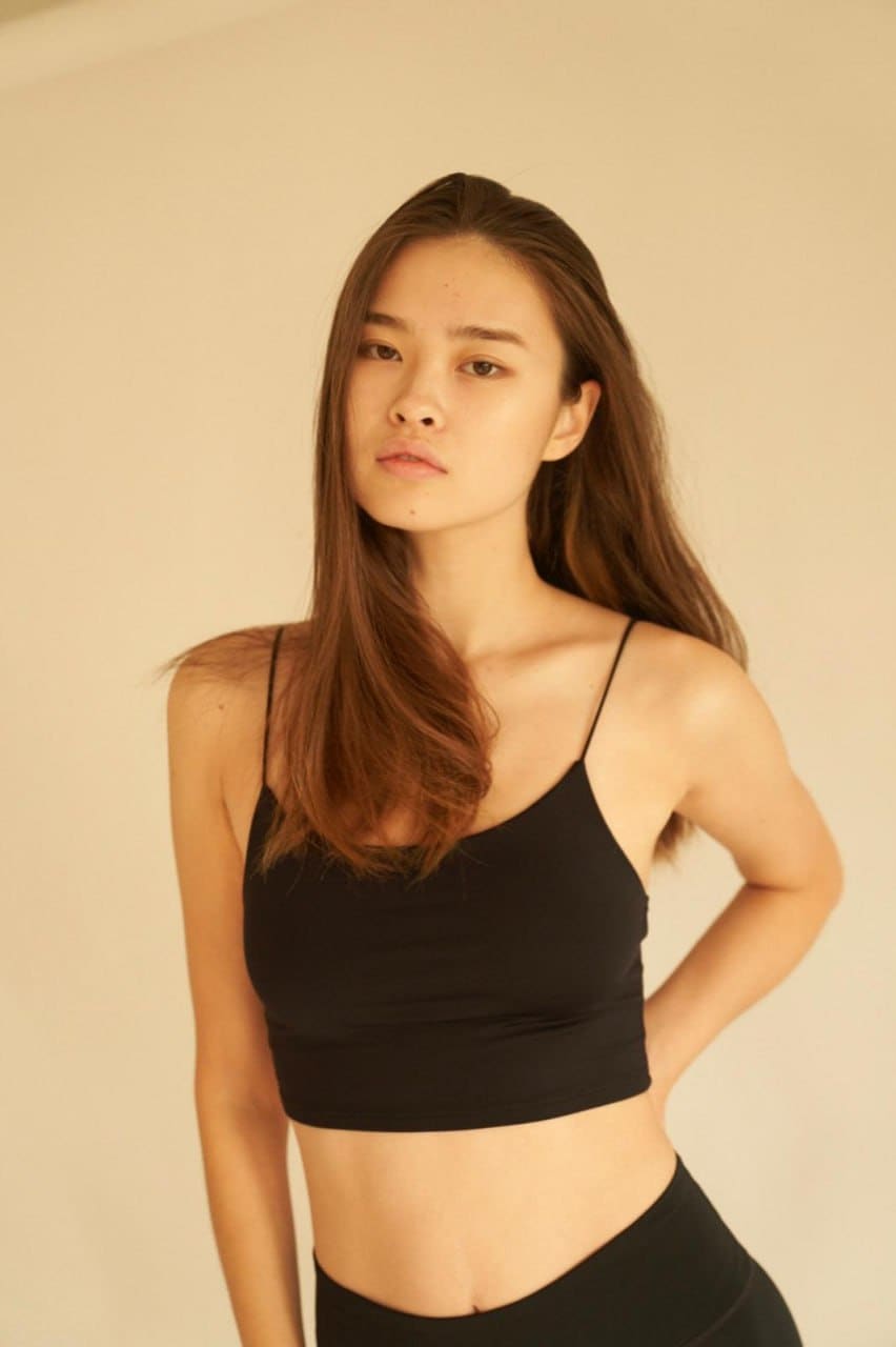 A young woman in a black crop top and high-waisted bottoms stands against a beige wall, one hand resting on her hip.