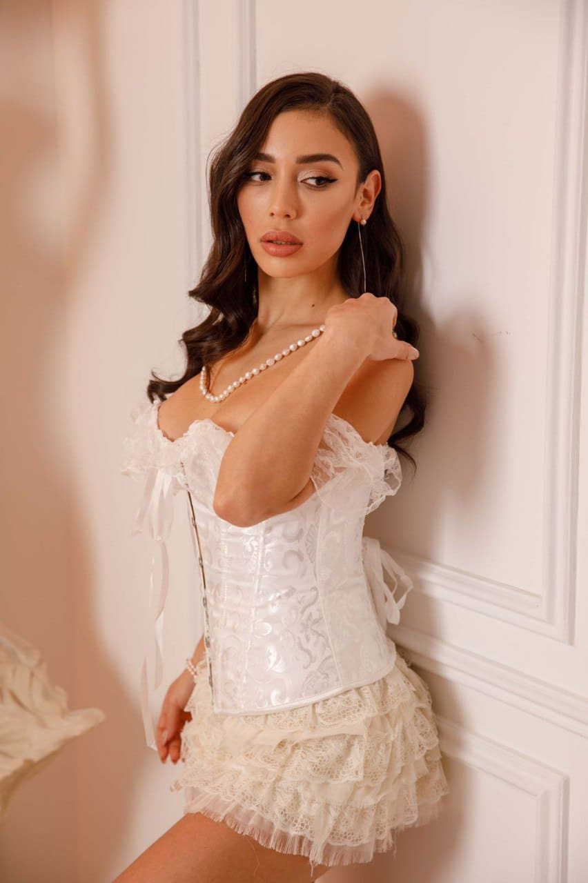 A woman stands by a white paneled wall in a white lace corset top with ruffles, matching skirt, and pearls, right hand raised near her shoulder.