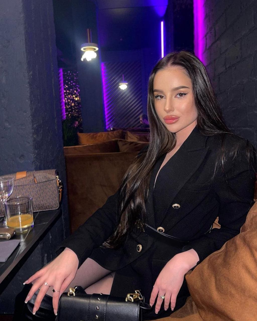 A woman in a black double-breasted blazer sits on a brown couch with a handbag on her lap and juice on the table.