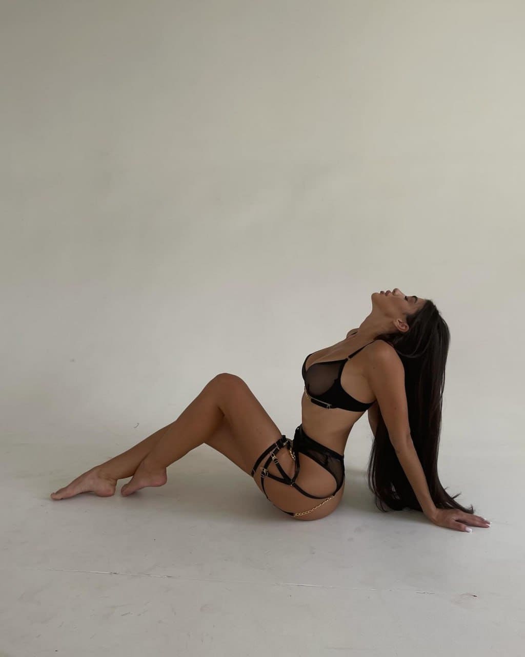 A woman sits on a light floor against a plain wall in black sheer lingerie with gold chain accents, head tilted back.