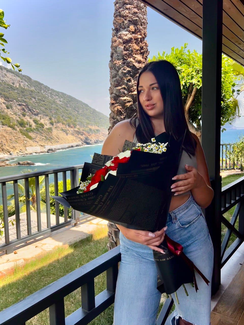 Mila stands on a balcony holding a bouquet wrapped in black paper, wearing a grey tank top and light blue jeans, with a coastal view behind her.