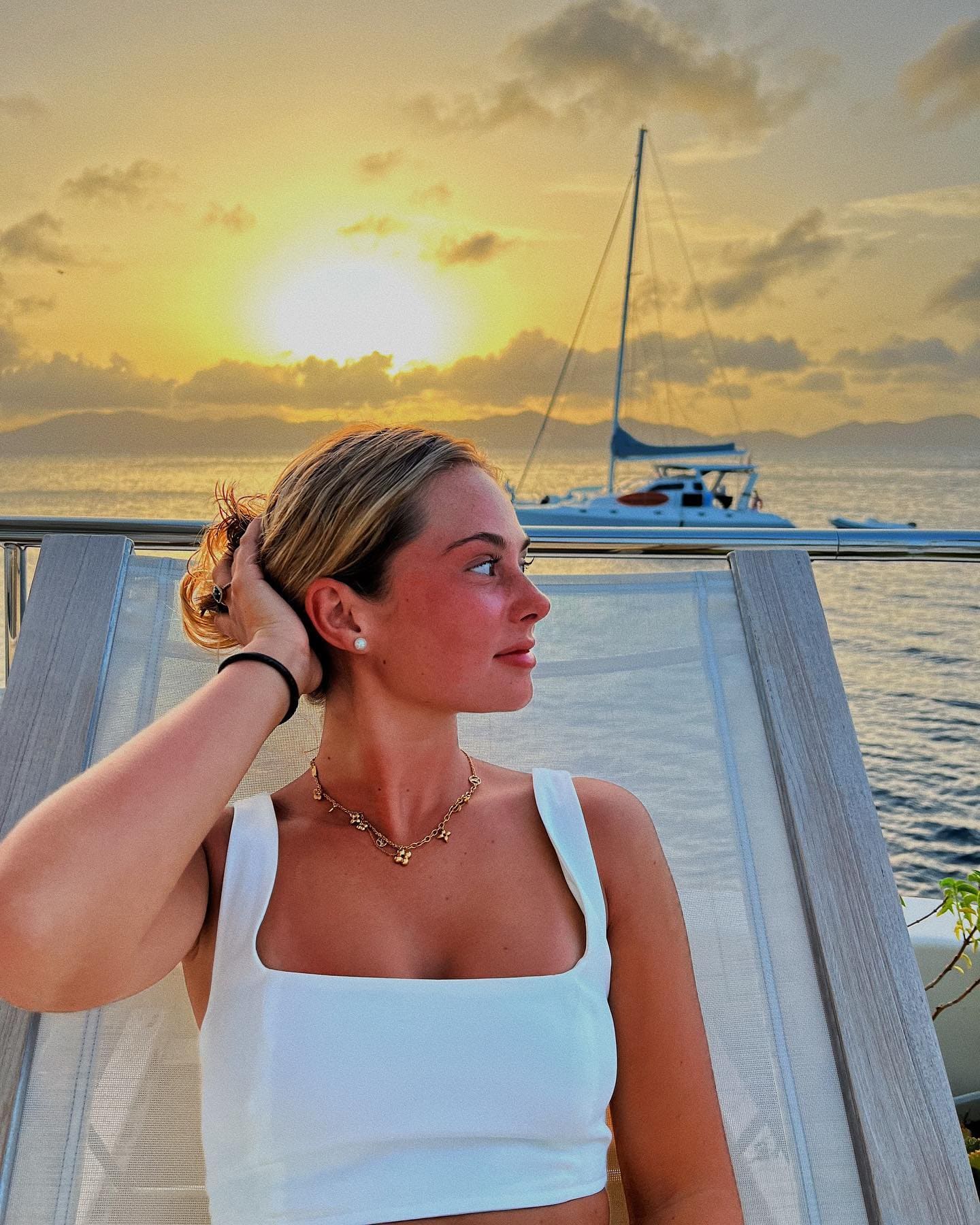 A woman stands on a boat deck at sunset in a white crop top and gold necklace, hand raised to her hair, with a sailboat in the background.