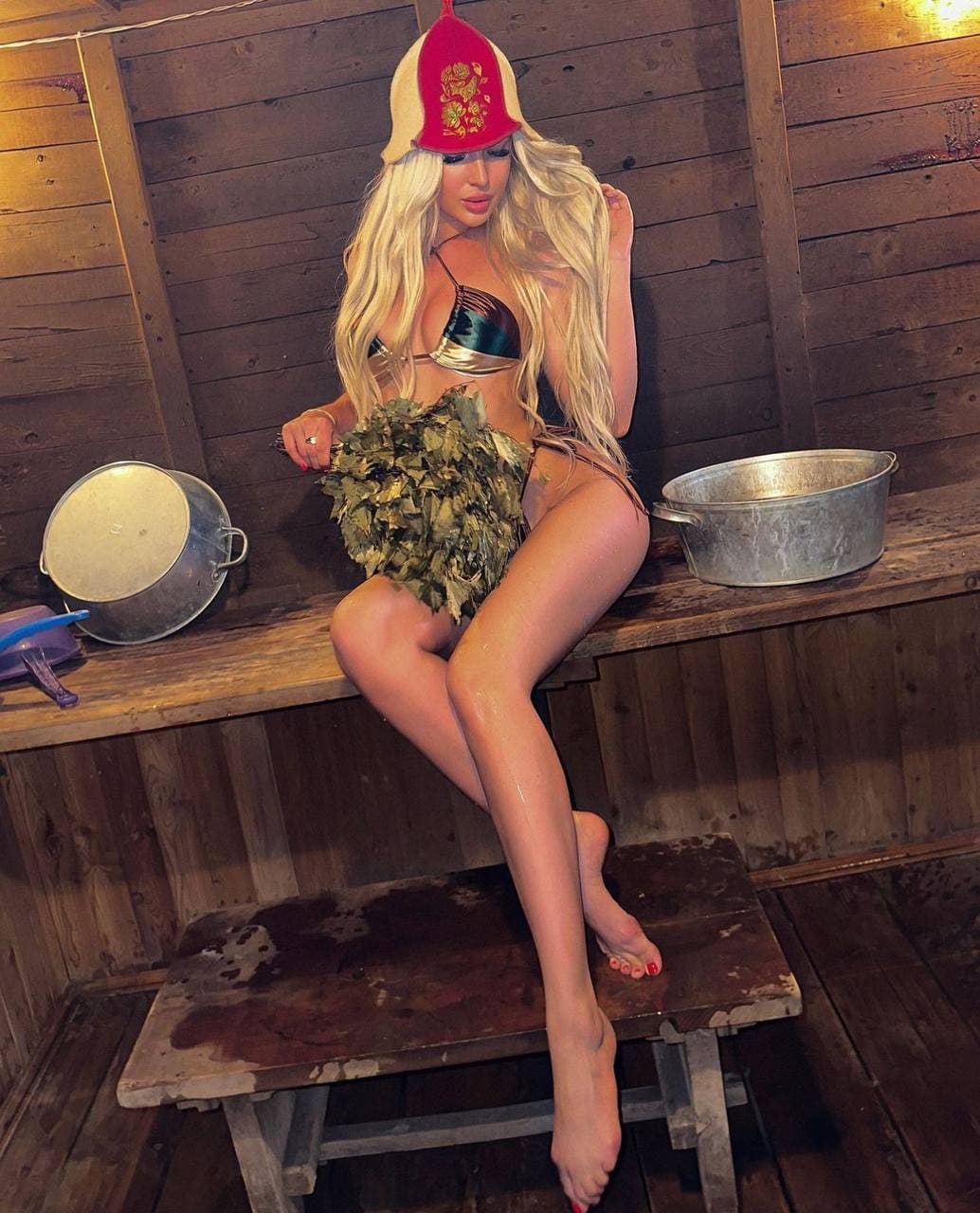 A blonde woman sits on a wooden bench in a rustic room wearing a red and gold hat, metallic bikini top, and leafy skirt, with metal pots behind.