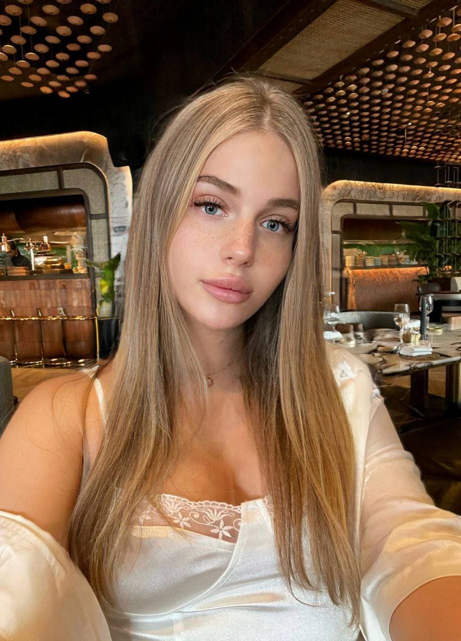 A freckled blonde woman takes a selfie in a restaurant, wearing a white satin camisole under a sheer blouse.