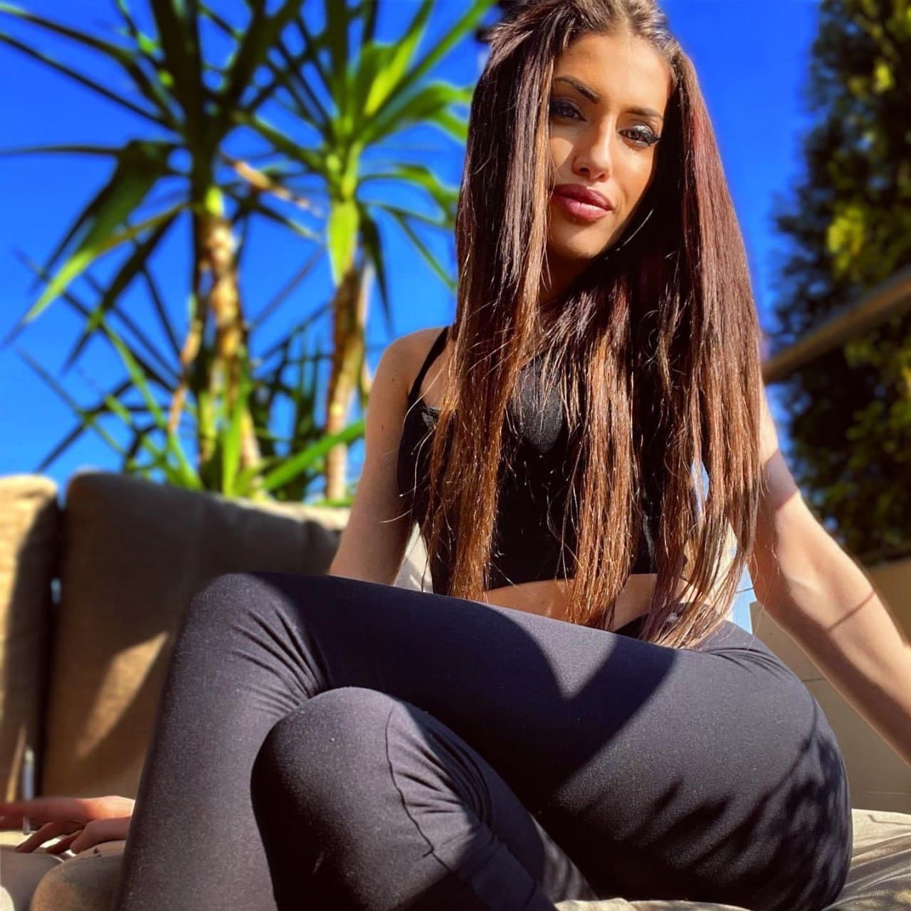 A woman in a black crop top and leggings sits on an outdoor bench with palm trees and a blue sky in the background.