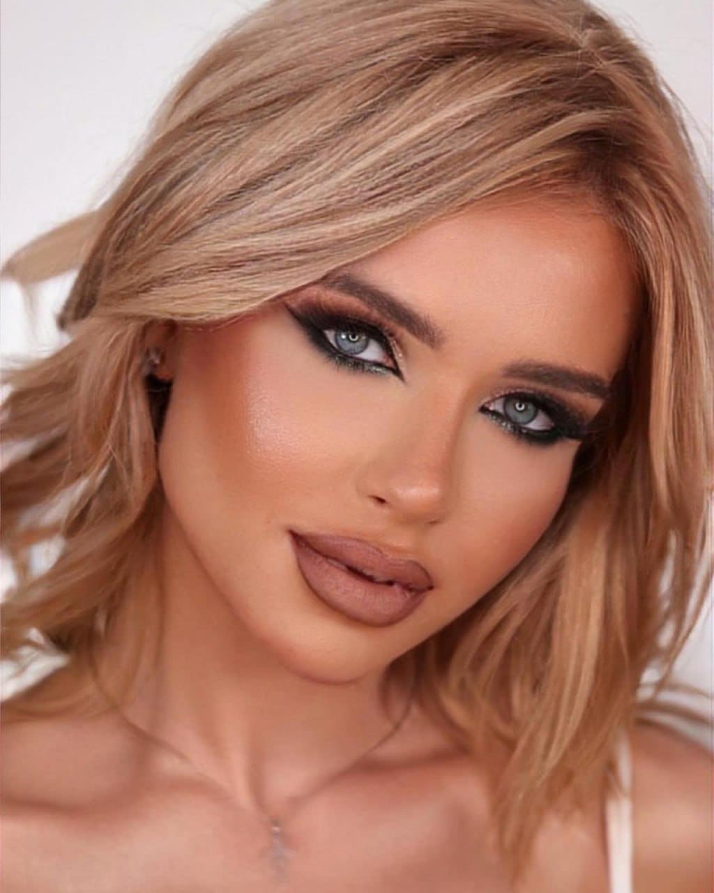 Close-up portrait of a woman with wavy blonde hair and dark eye makeup, wearing a thin-strapped top against a light background.