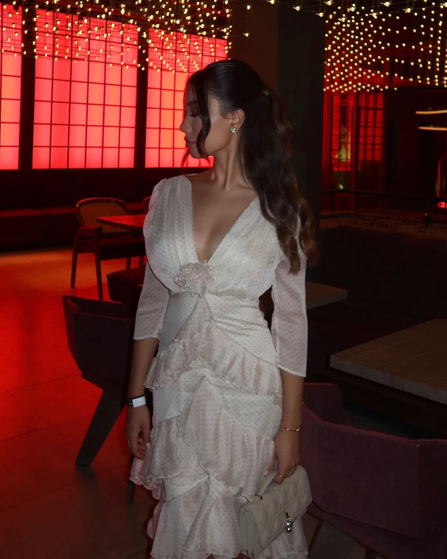 A woman in a white polka-dot dress stands in profile holding a quilted bag, with red-lit windows and string lights.