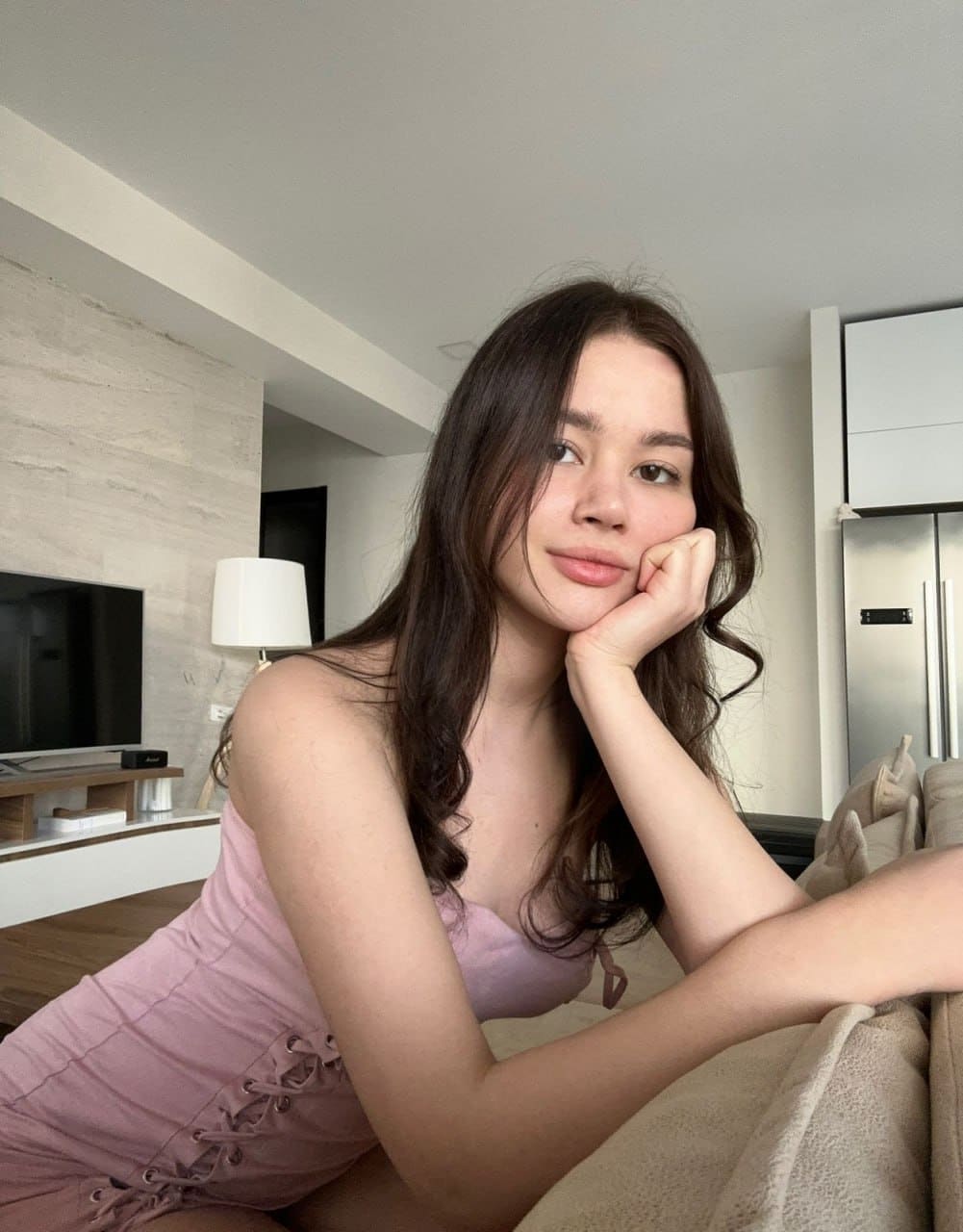 A woman in a sleeveless pink lace-up dress sits on a beige couch, resting her chin on her hand in a modern living room.