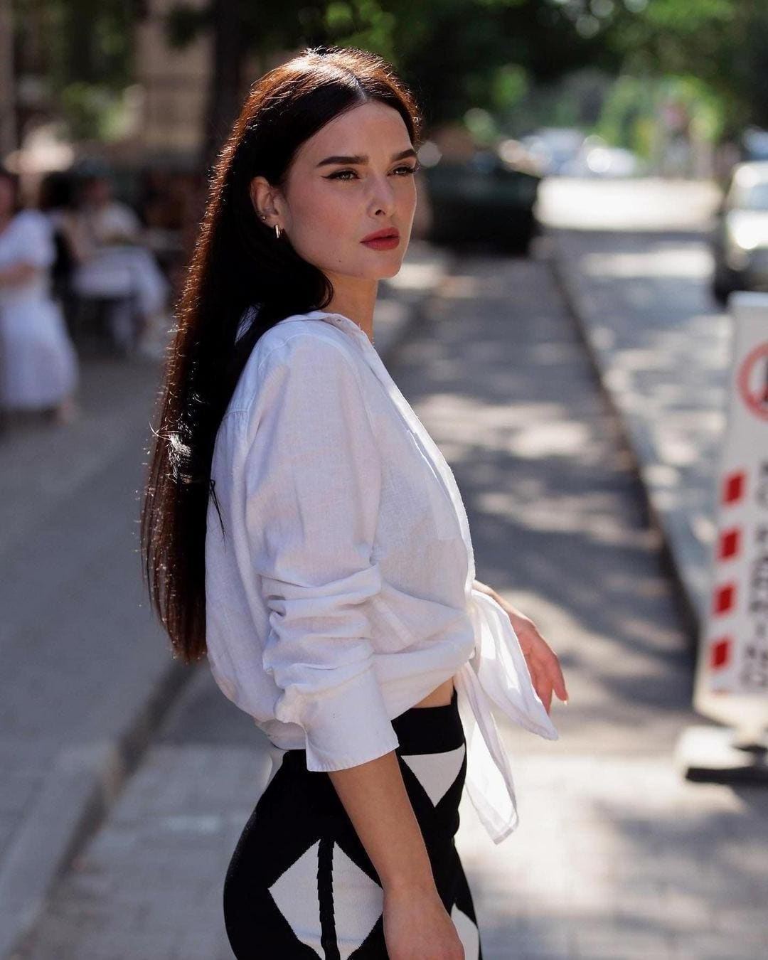 Belle stands on a sunlit city sidewalk in a white tied shirt and geometric skirt, looking back over her shoulder.