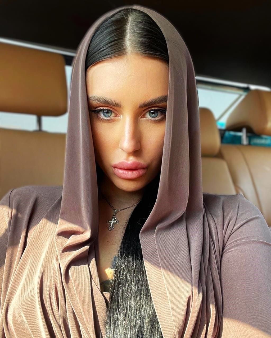 A woman with blue eyes sits in a car with tan leather seats, wearing a mauve hooded top and cross necklace.