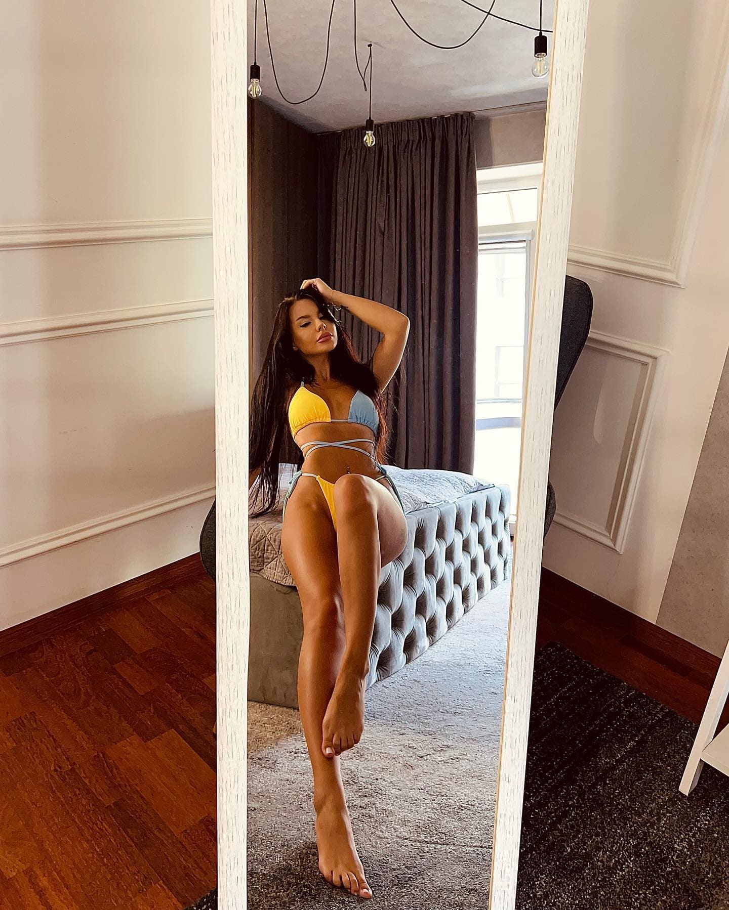 A woman stands barefoot in a bedroom, posing in a yellow and blue string bikini before a mirror with one hand raised to her hair.