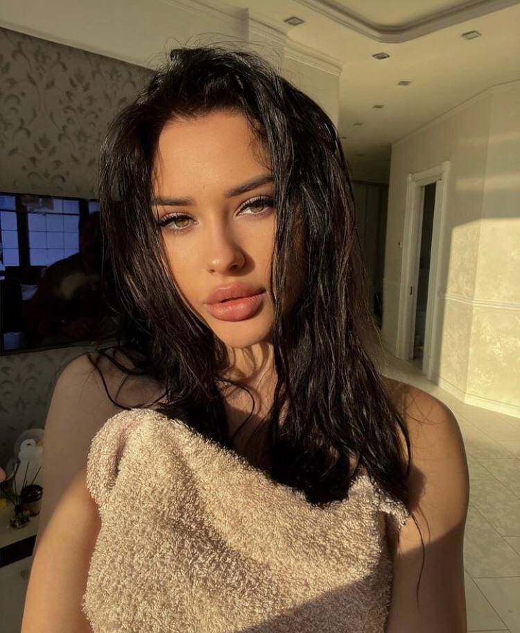 Vlada stands in a sunlit room, wrapped in a beige towel, with wet dark hair and bare shoulders.