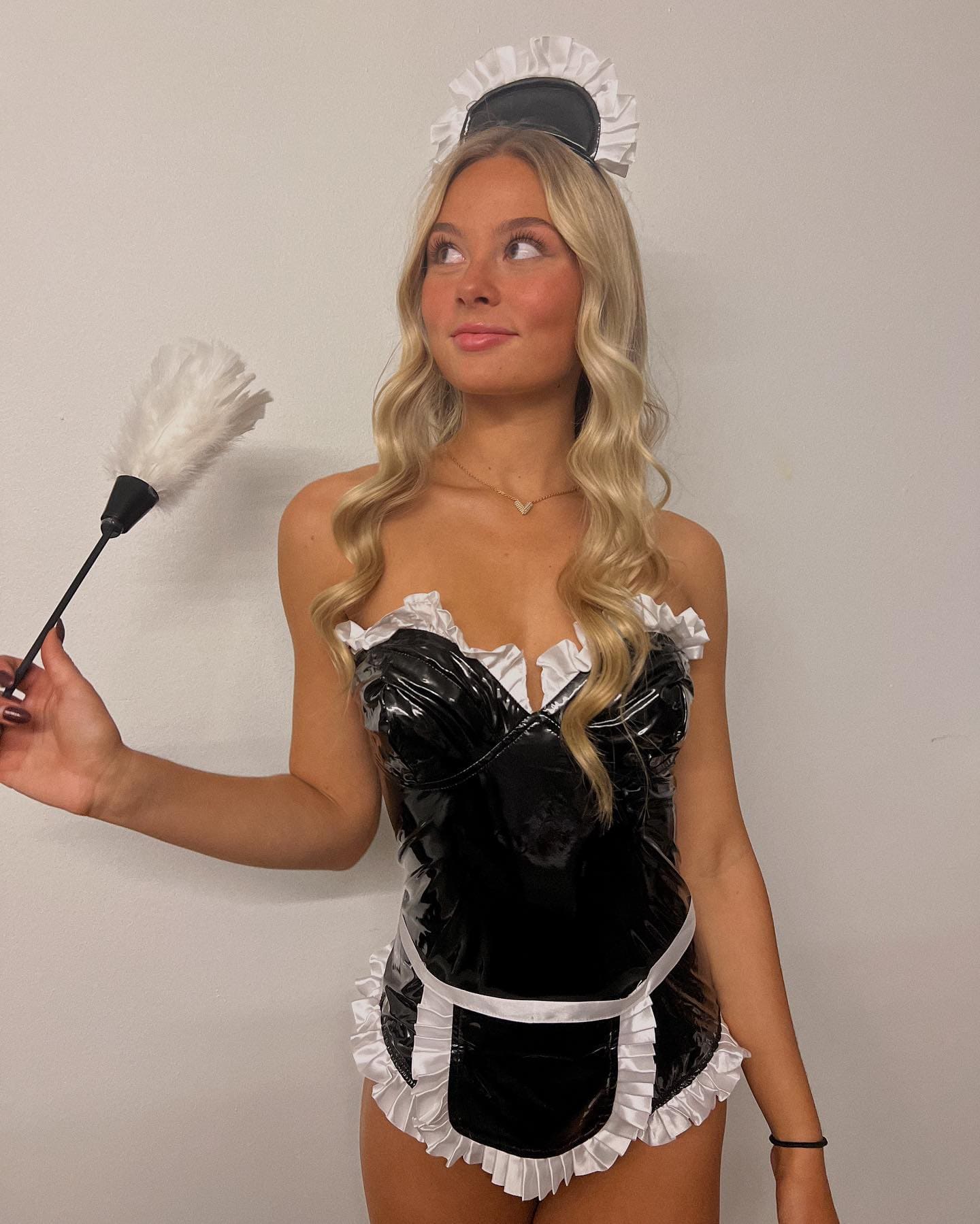 A woman in a black and white maid costume with a ruffled headband holds a feather duster while standing against a plain wall.