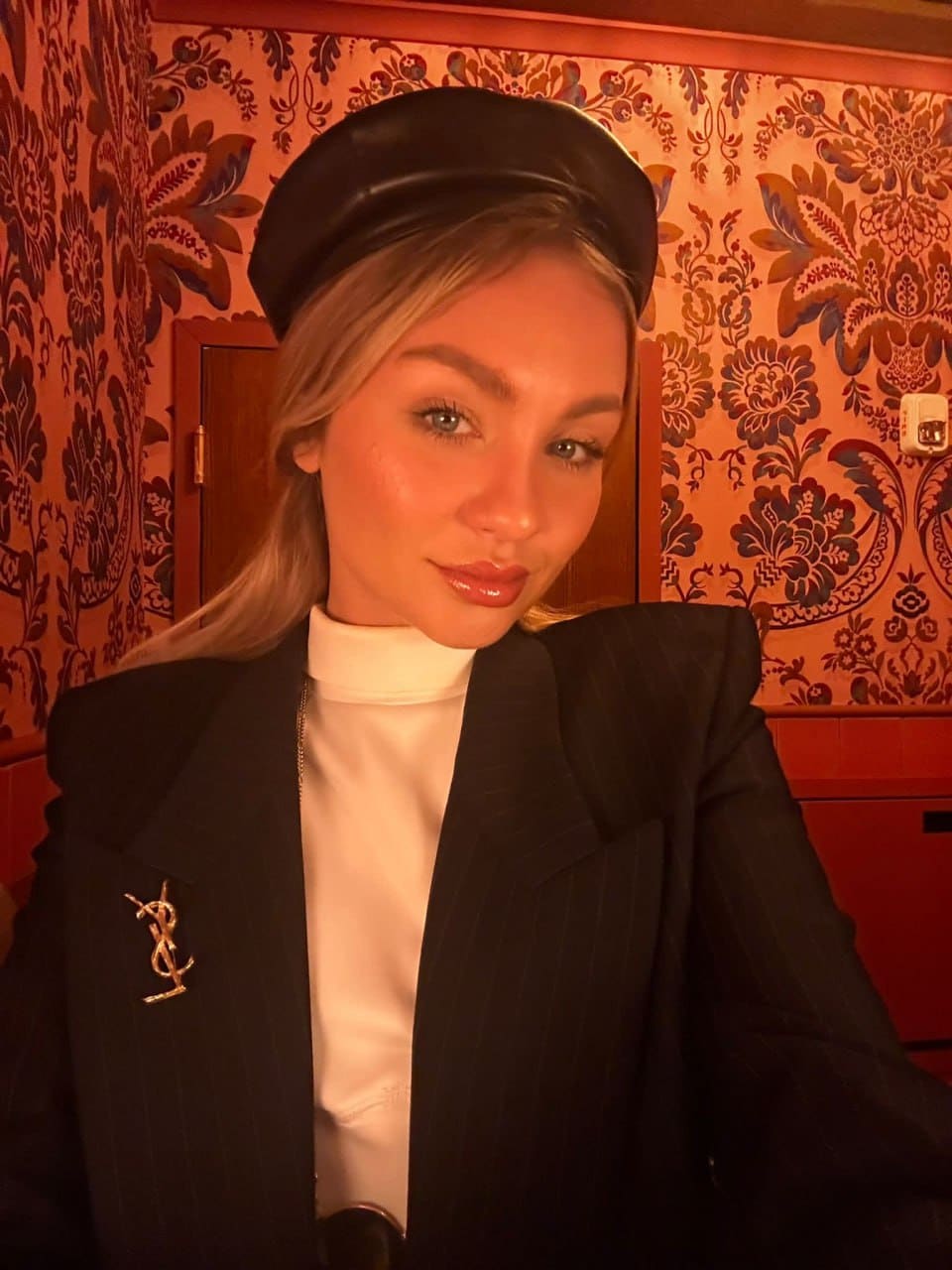 A blonde woman wears a black leather beret, white turtleneck, and dark pinstriped blazer with a gold brooch, posing in front of a floral patterned wall.