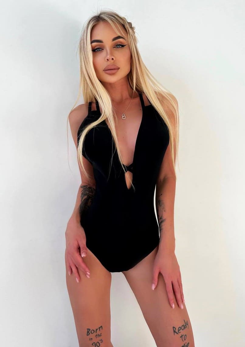 A woman with long blonde hair and tattoos stands against a white wall in a black cut-out swimsuit, hands resting on her upper thighs.